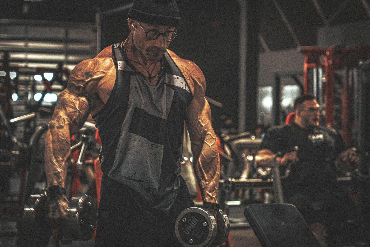 Tattooed bodybuilder in a beanie and round glasses holding dumbbells on a gym floor