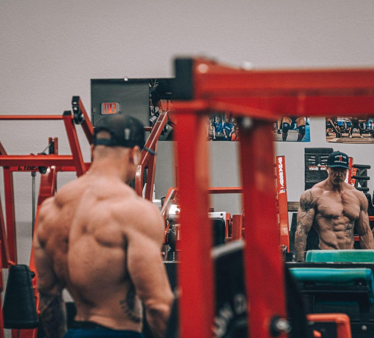 Shirtless bodybuilder framed by a red rack, his reflection facing the camera