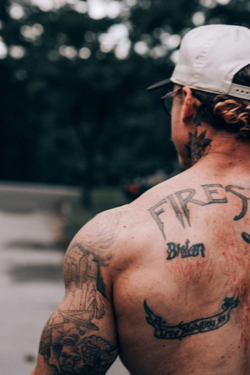 Tattooed back and shoulder of an athlete outdoors, trees blurred behind