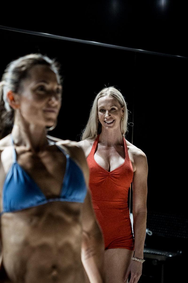 Posing coach smiling in the mirror behind a bikini competitor