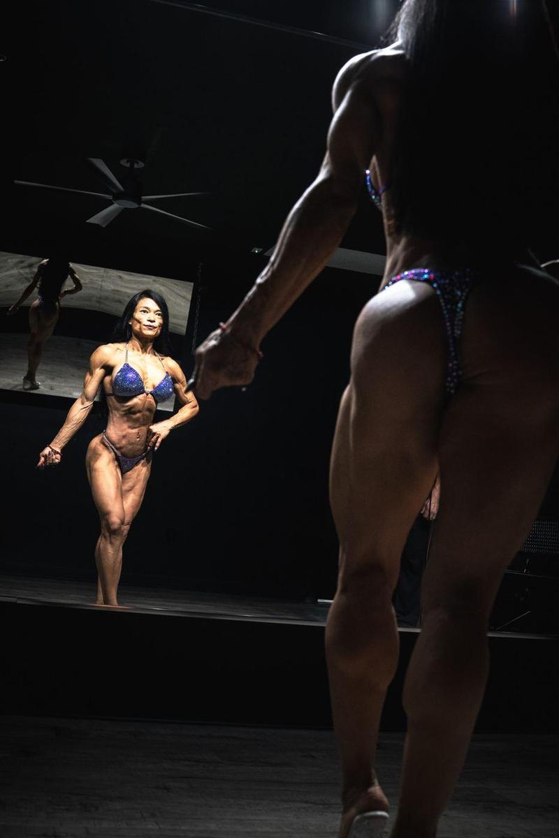Figure competitor posing toward the mirror under a single spotlight